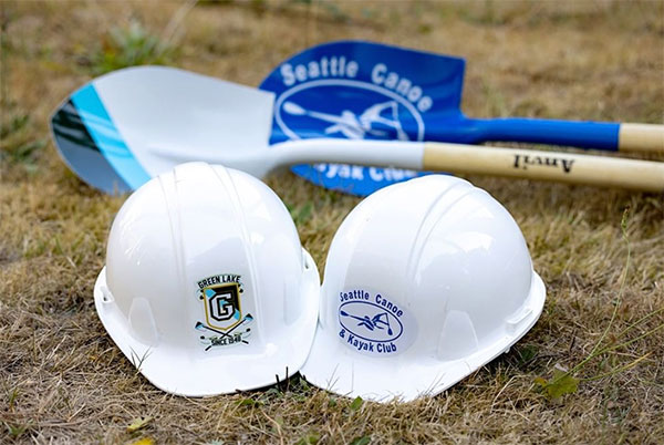 Two construction hardhats sit on grass, one has a Green Lake Crew logo and the other has a Seattle Canoe and Kayak Club logo. Behind the hardhats are two shovels, one painted like a GLC oar and the other with SCKC's logo.